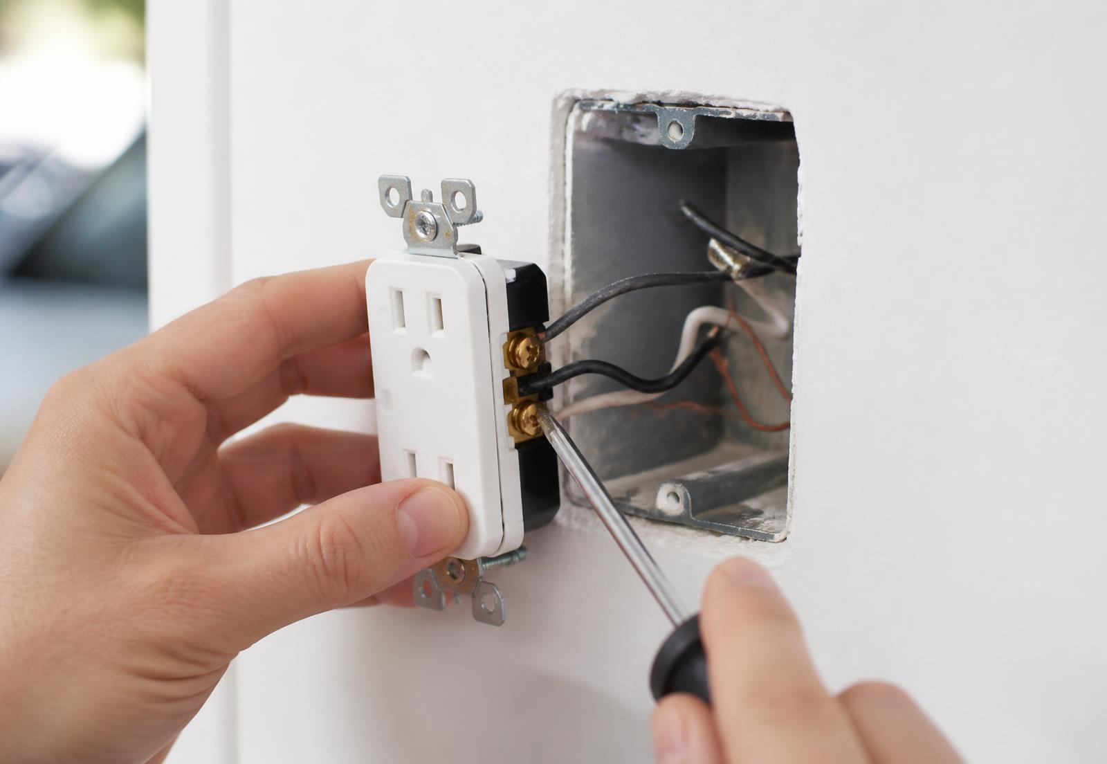 Hands installing a new white electrical receptacle into a wall box with wiring visible